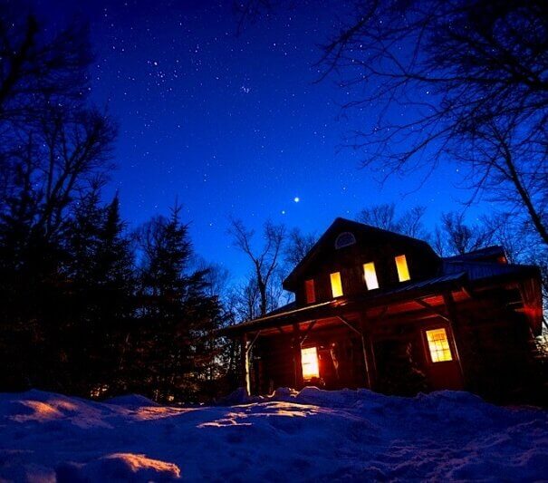 Algonquin Log Cabin during night time with a sky full of stars and covered in snow