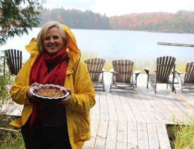 Woman standing outside on a deck with Muskoka chairs, with in the background a lake and beautiful Fall colours